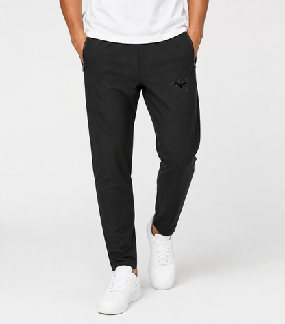 A person wearing slim black jogger-style pants with a small black embroidered logo on the upper left thigh, paired with a white t-shirt and white sneakers, photographed against a clean light gray studio background.
