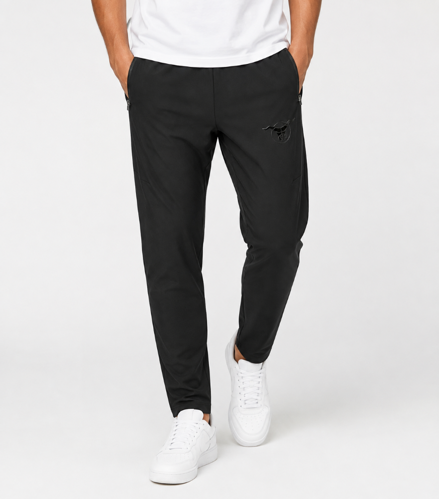 A person wearing slim black jogger-style pants with a small black embroidered logo on the upper left thigh, paired with a white t-shirt and white sneakers, photographed against a clean light gray studio background.