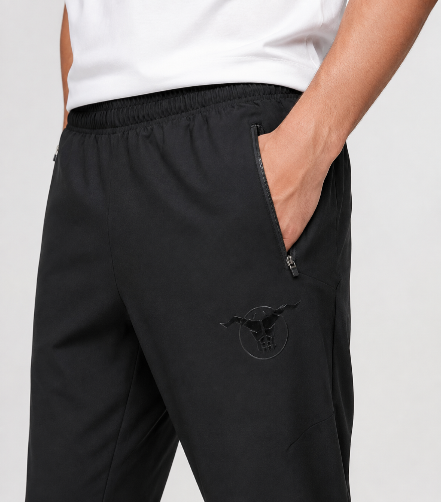 Close-up of a person wearing black athletic pants with a side zip pocket and a tonal bull logo on the thigh, paired with a white top against a light gray background.